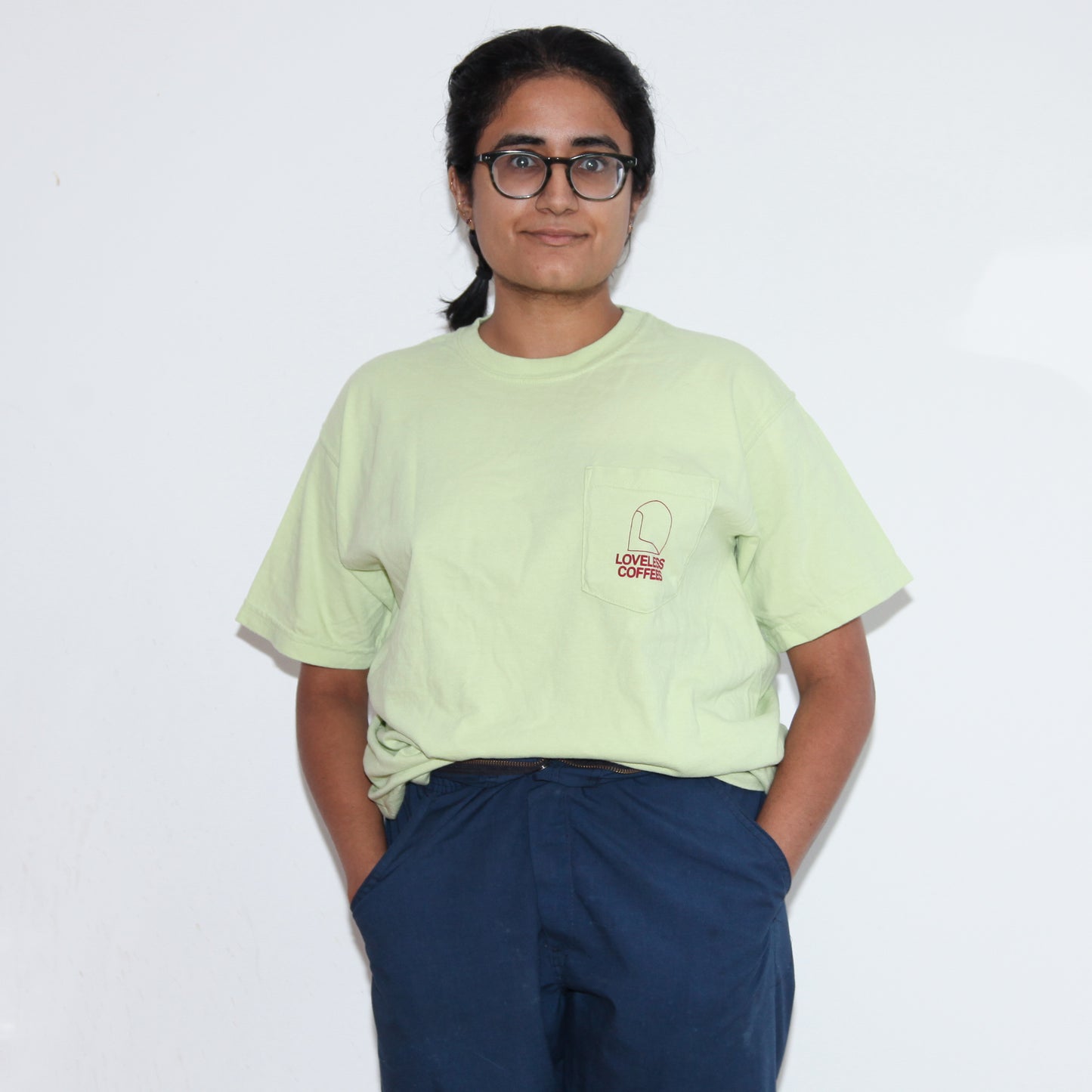 A woman wearing a light green pocket tee with 'LOVELESS COFFEES' text in red on the pocket