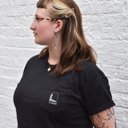 A person wearing a black t-shirt with a pocket, and a logo on the chest, standing in front of a white brick wall.