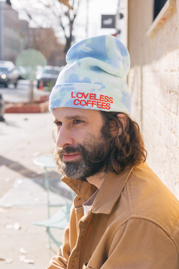 A person wearing a blue & green tie-dye beanie with 'LOVELESS COFFEE' embroidered on it.